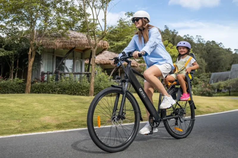 A woman wearing a helmet rides a bicycle with a young child, also in a helmet, seated in a rear child seat on a sunny day. They are passing green lawns and houses with thatched roofs.