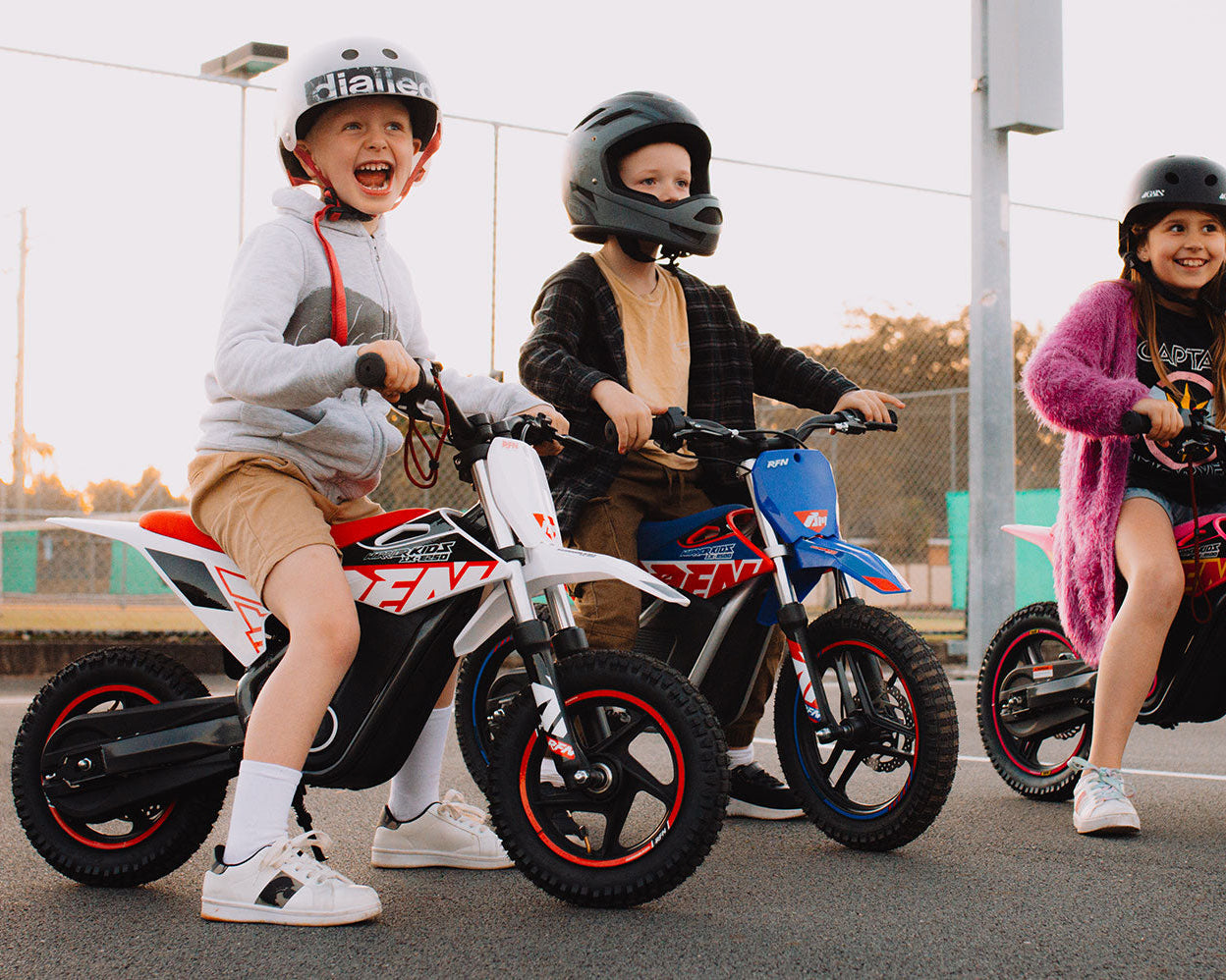 Three children wearing helmets riding small electric dirt bikes on a paved surface outdoors at sunset