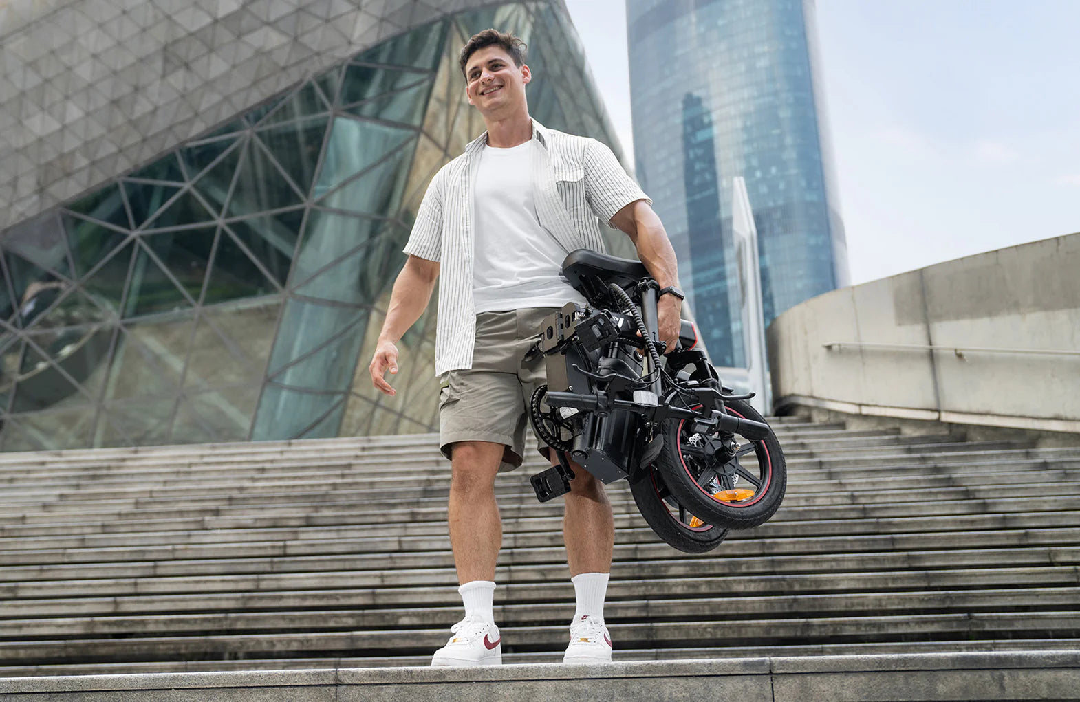 A man in casual clothing smiles while carrying a folded black bicycle up outdoor steps, with modern glass buildings in the background.