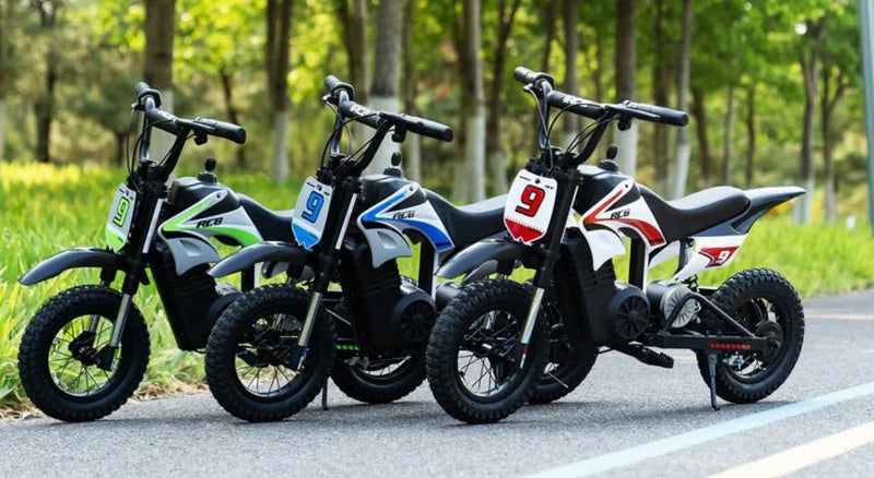 Three small electric dirt bikes with number plates are parked on a paved path outdoors, surrounded by green grass and trees on a sunny day.
