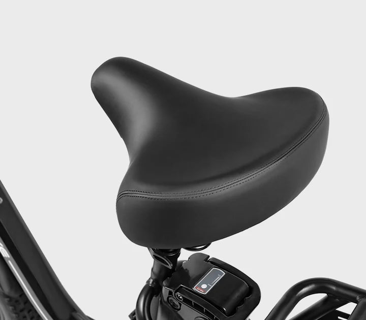 A close-up view of a black, wide, cushioned bicycle seat attached to a bike frame, with part of the handlebar and an electronic display visible below the seat, against a plain light gray background.