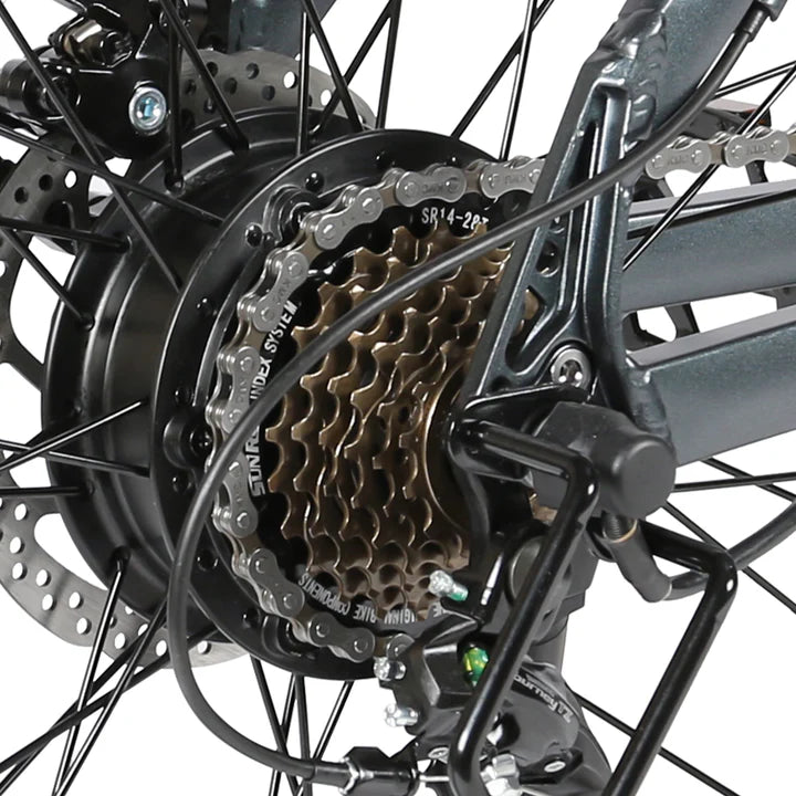 Close-up of a bicycle rear wheel showing the gear cassette, chain, derailleur, and disc brake rotor. The components are metallic with black and bronze tones, highlighting the bikes drivetrain and braking system.
