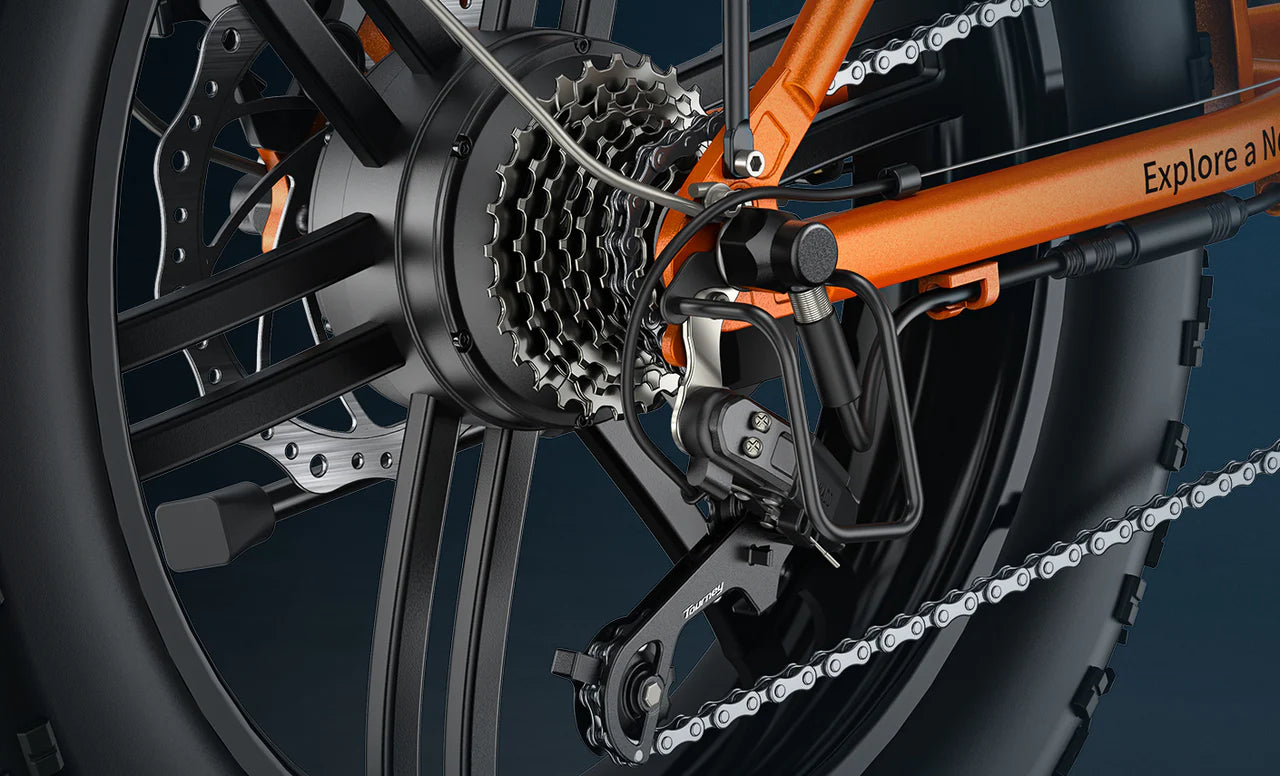 Close-up of the rear wheel and gear system of an orange bicycle, showing the gears, derailleur, chain, and part of the frame with Explore a N text visible.
