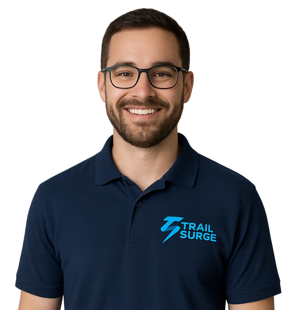 A smiling man with short brown hair, beard, and glasses is wearing a navy blue polo shirt with the TRAIL SURGE logo in blue text on the chest, standing against a plain white background.