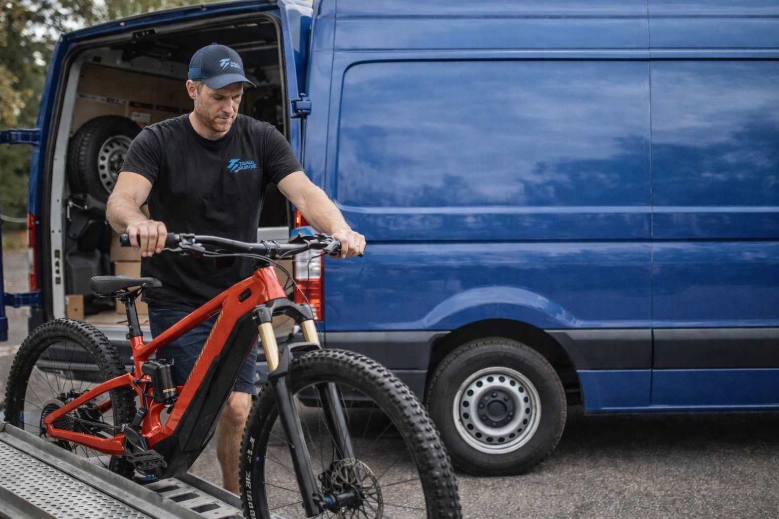 Man wearing a black cap and shirt pushing a red mountain bike up a ramp into a blue van outdoors.