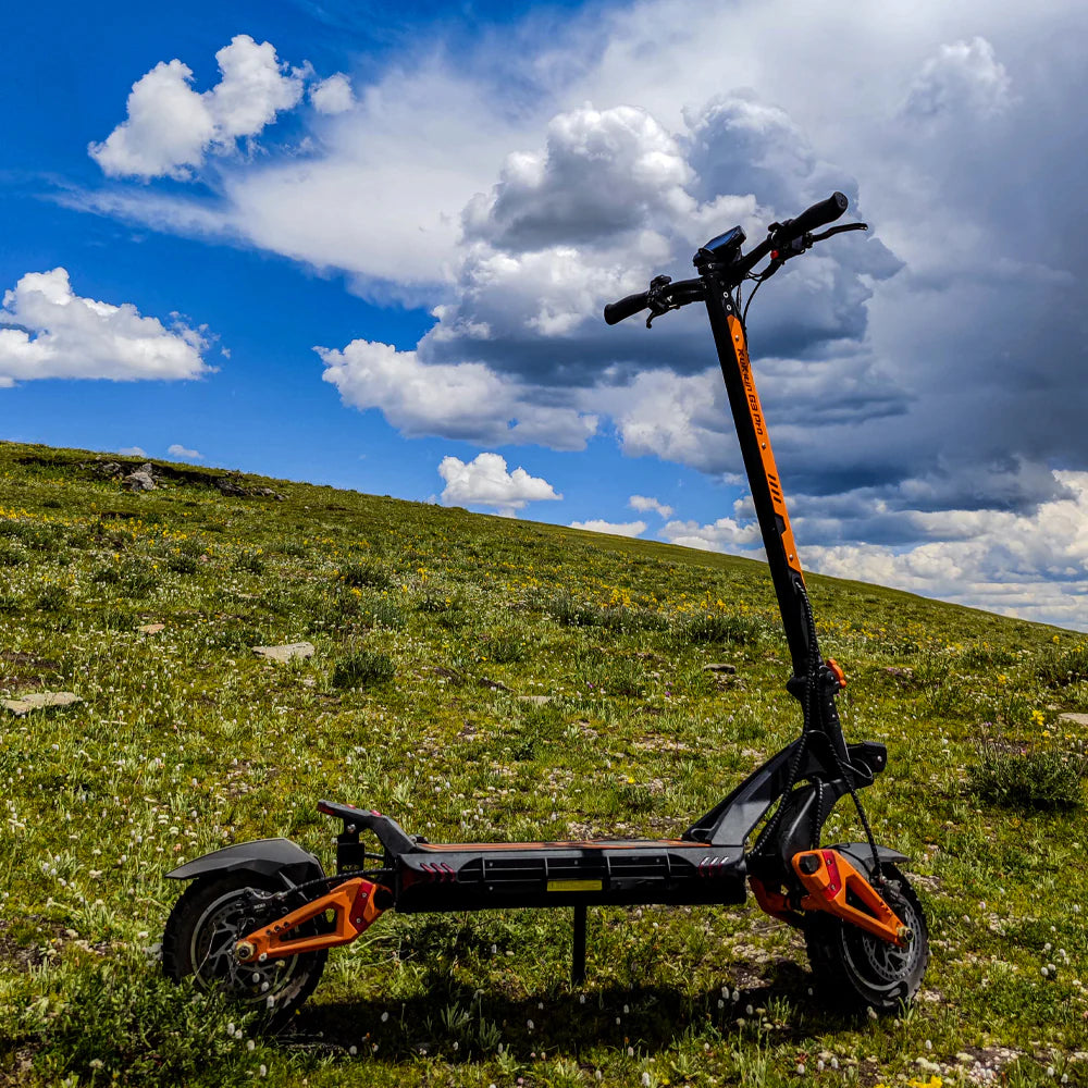 Black and orange electric scooter standing on grassy hill under a partly cloudy blue sky