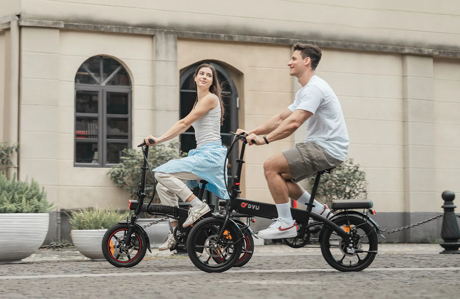 A woman and a man smile while riding black electric bicycles on a paved street, with a beige building and potted plants in the background.