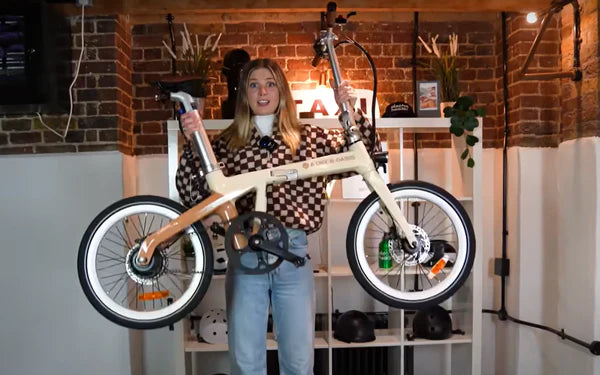 A woman wearing a checkered sweater and jeans holds up a beige folding bicycle indoors, standing in front of shelves with plants, helmets, and bottles against a brick wall.