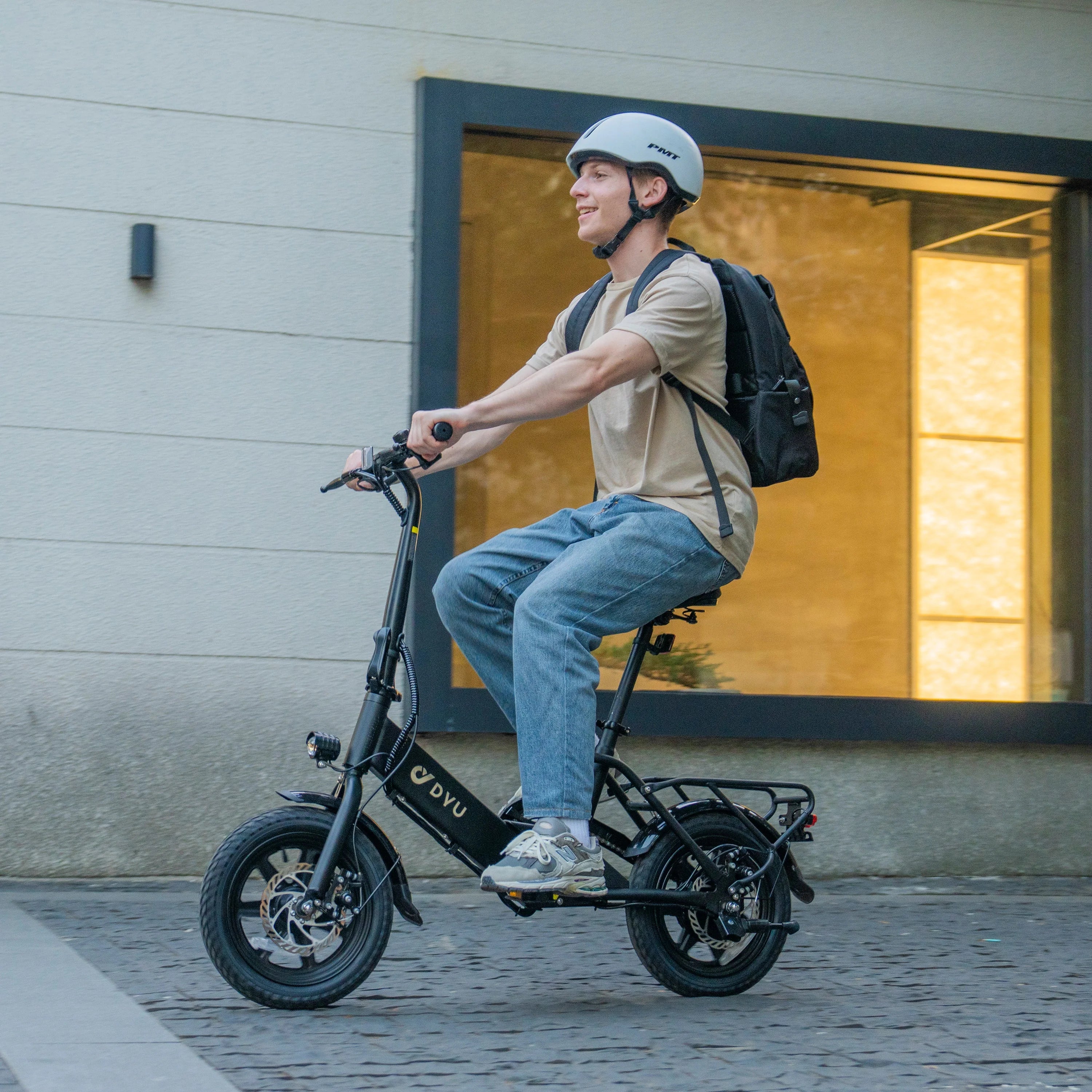 A young man wearing a helmet and backpack rides the DYU C3 Mini Electric Bike with a 250W rear motor on a cobblestone street in front of a modern building with large windows.