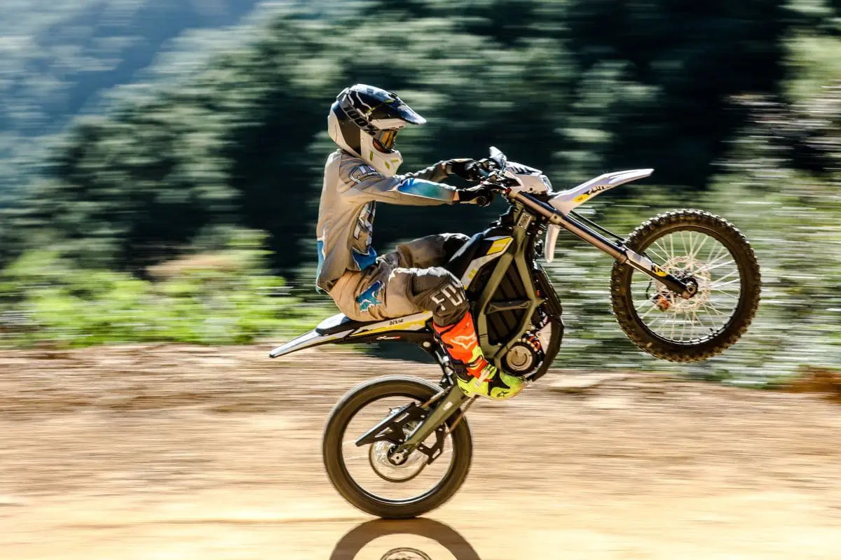 Motorcyclist wearing protective gear performing a wheelie on a dirt trail with blurred forest background