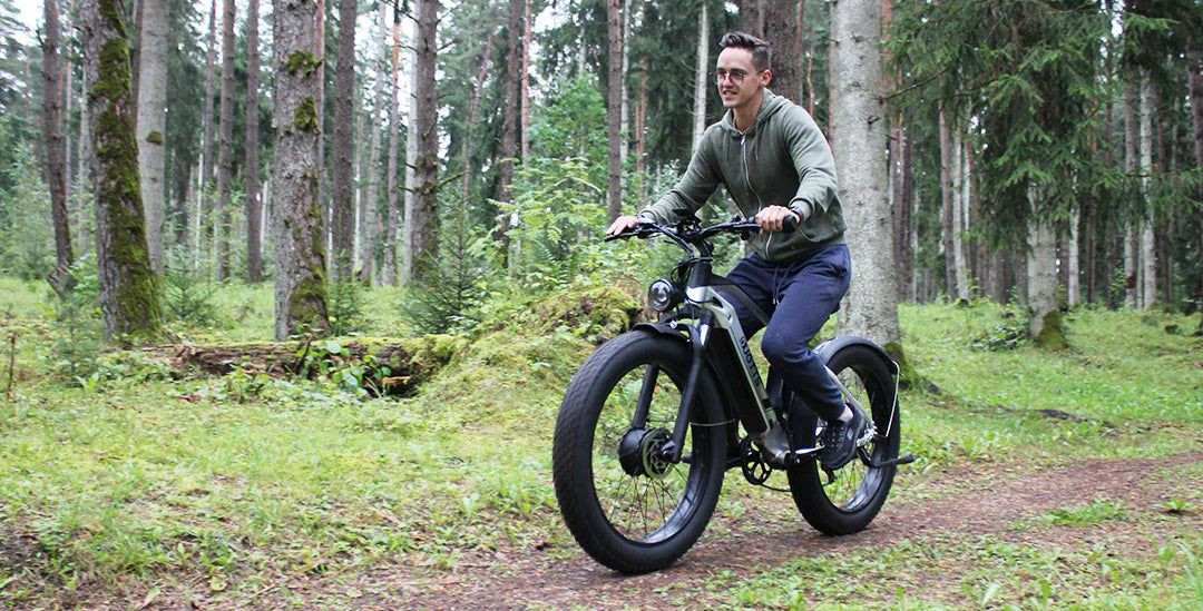 A person rides a fat-tire electric bike along a dirt path through a lush, green forest, surrounded by tall trees and mossy ground.