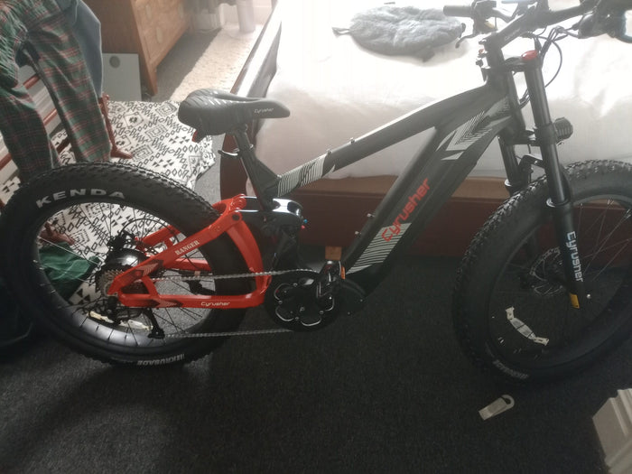 A black and red electric mountain bike with thick tires labeled KENDA is parked indoors on a dark carpet, near a bed and some furniture.