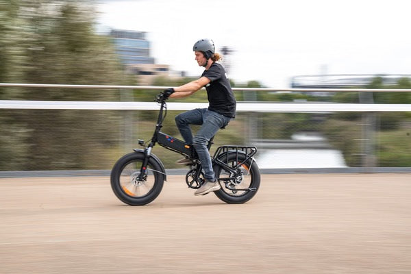 A person wearing a helmet rides a black electric bike quickly along a path, with background blurred to show motion. Trees and buildings are visible in the distance.