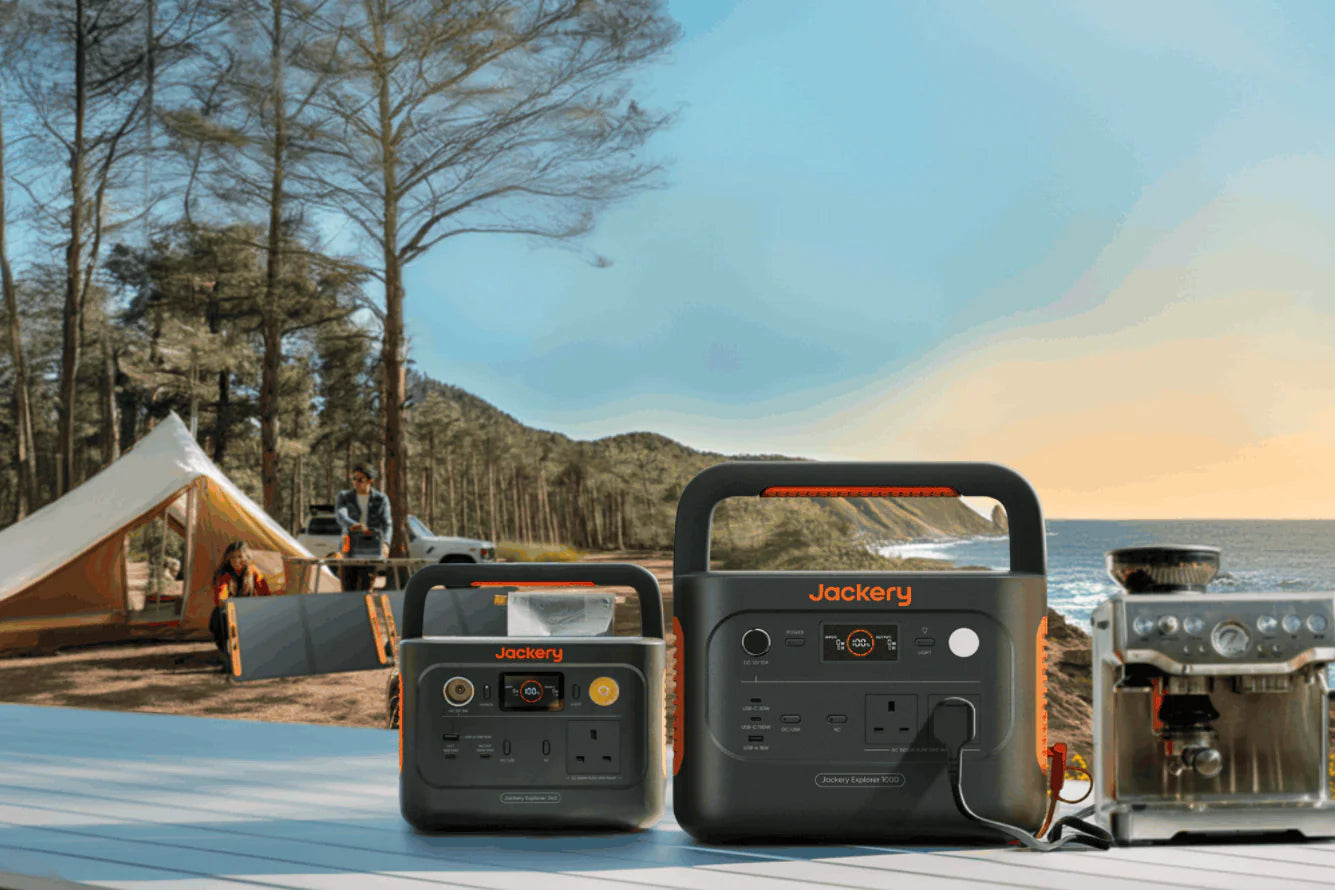Two Jackery portable power stations on a table with people camping and a scenic lake at sunset in the background