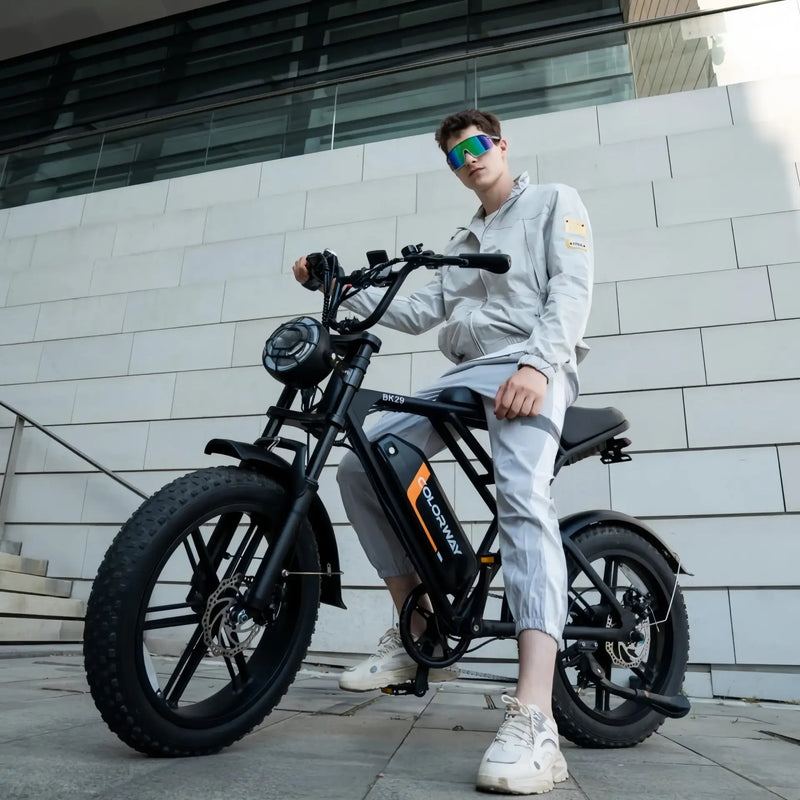 A young man in light gray clothing and reflective sunglasses poses confidently on a black e-bike in front of a modern white-tiled building.