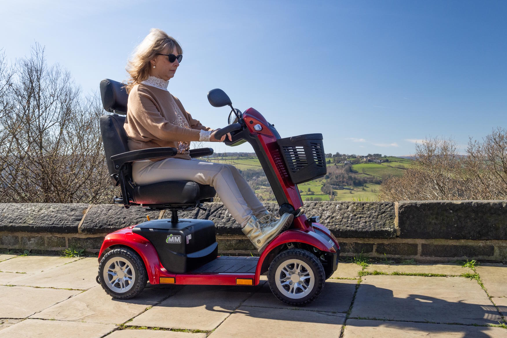Woman wearing sunglasses riding a red mobility scooter on a paved path with a rural landscape background