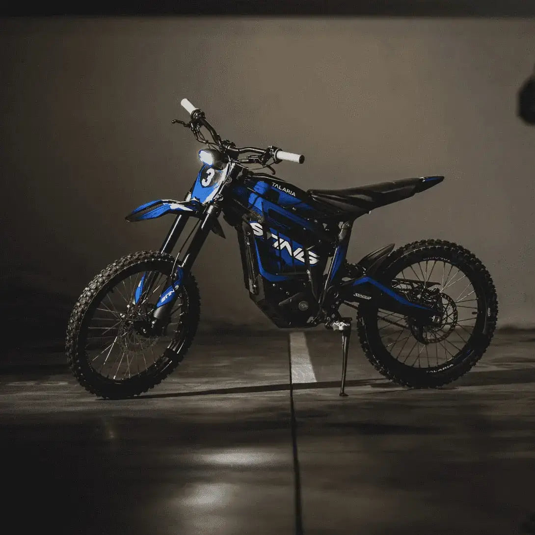 Blue and black electric dirt bike with knobby tires and white handlebar grips on a dark concrete floor background