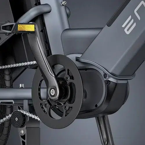 Close-up view of an electric bicycles crankset and pedal area, showing the chain, pedal, and battery compartment, with a modern, sleek black and gray design.