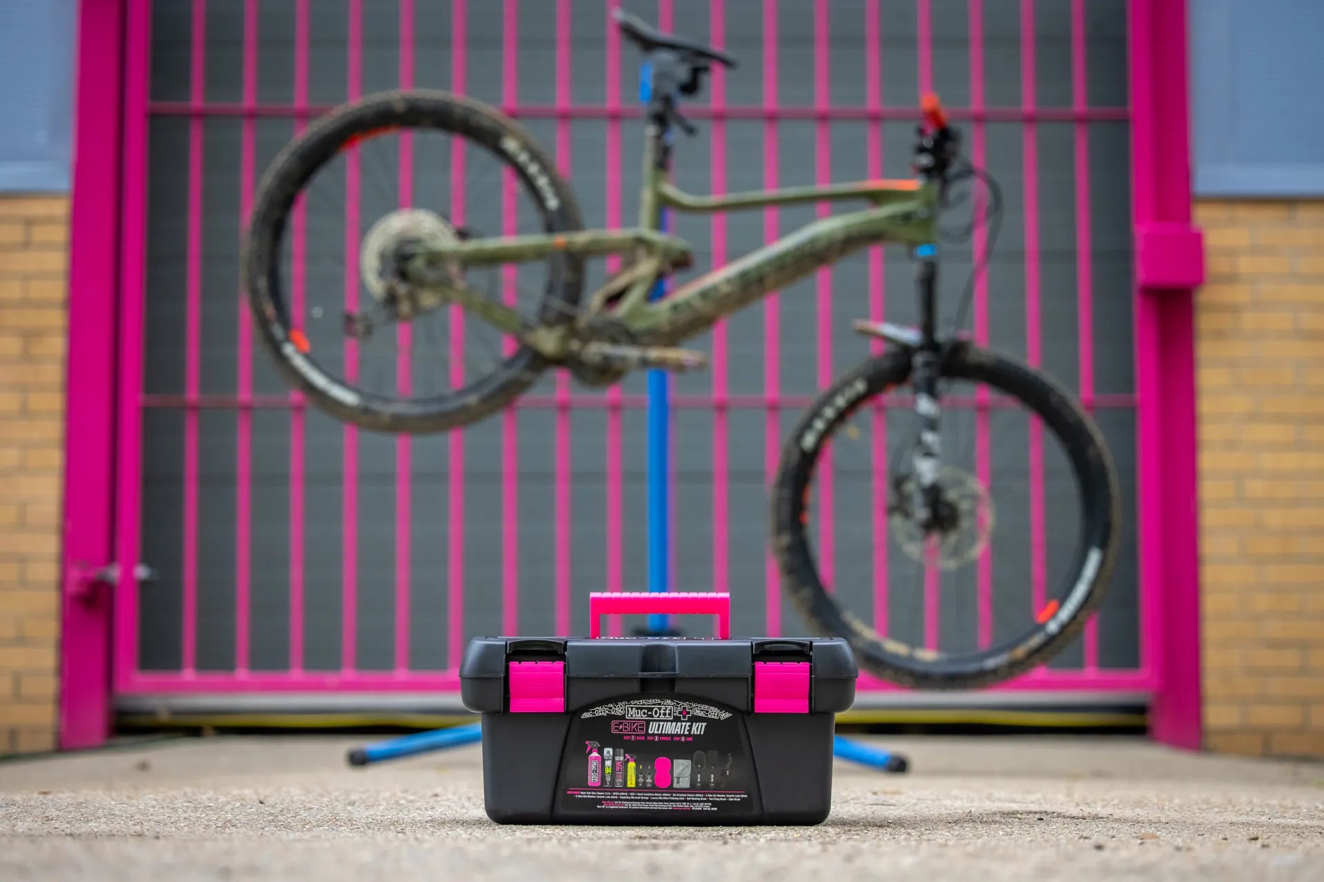 Black and pink toolbox on the ground with a dirt-covered mountain bike suspended in the background outdoors