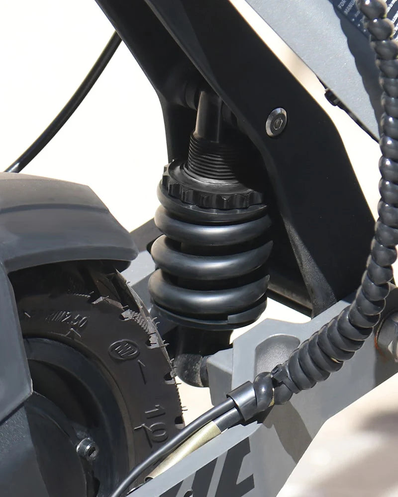 Close-up of the Vipcoo VIPCOO VS6 1000W Electric Scooter's black and silver rear suspension, featuring a coiled spring, wheel with visible tire tread, and wiring in the foreground.