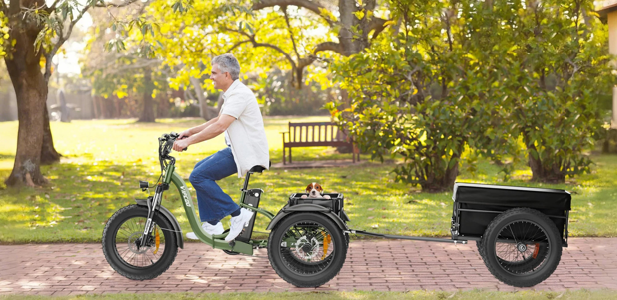 A man rides an electric bike with a small trailer on a park path. A small dog sits in a carrier behind him. Green trees and sunlight fill the background, creating a peaceful outdoor scene.