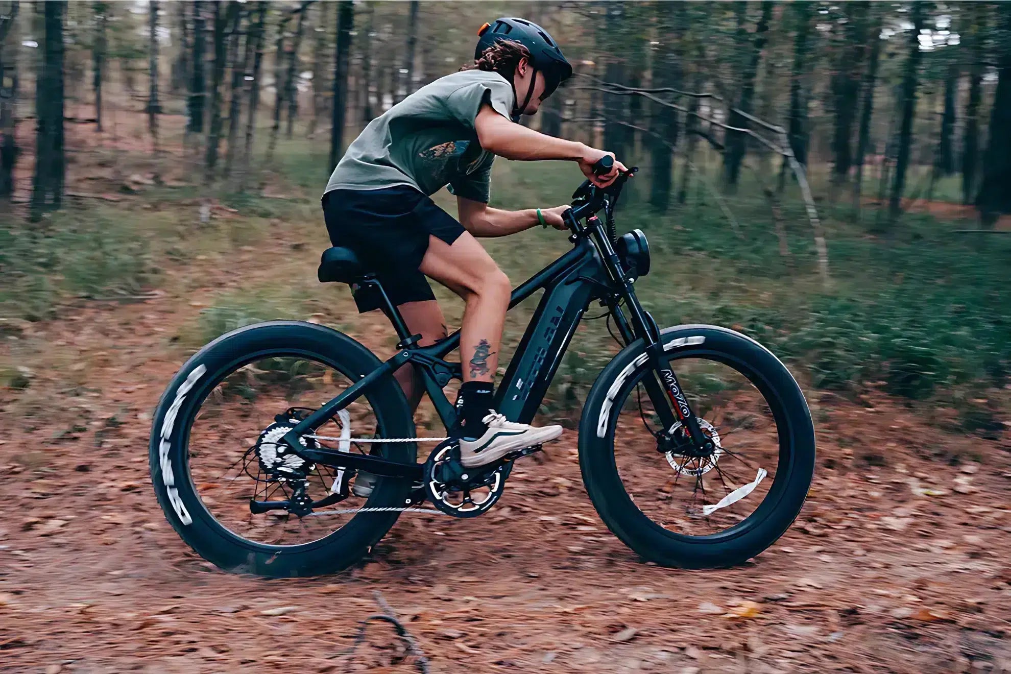 Young person wearing a helmet riding a black electric mountain bike on a forest trail in autumn.