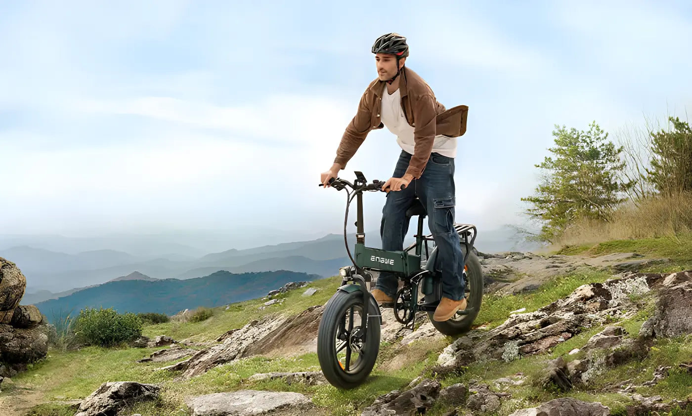 A man wearing a helmet and casual clothes rides an electric bike on a rocky trail with mountains and a cloudy sky in the background.