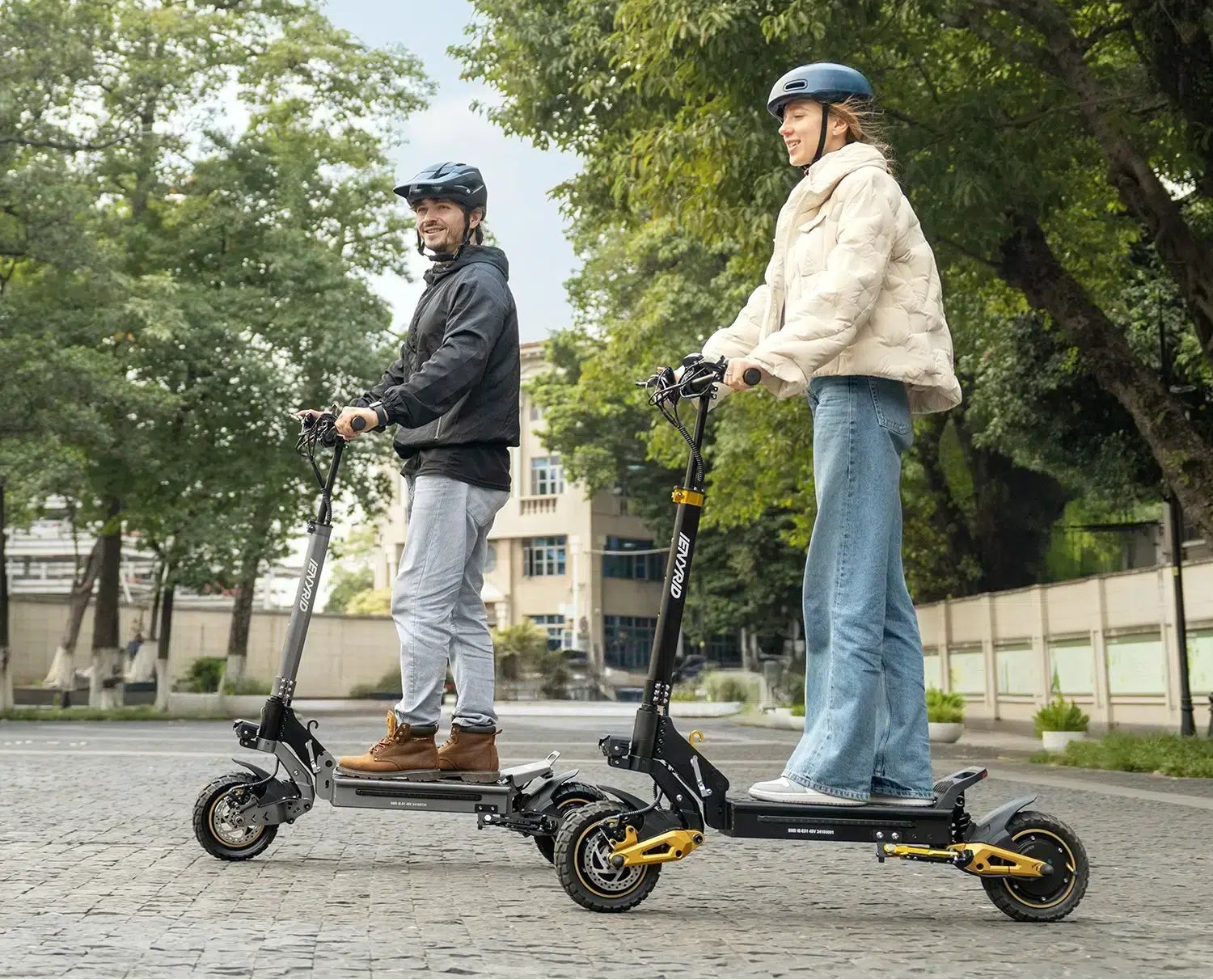 Two people wearing helmets riding electric scooters on a paved path with trees and buildings in the background