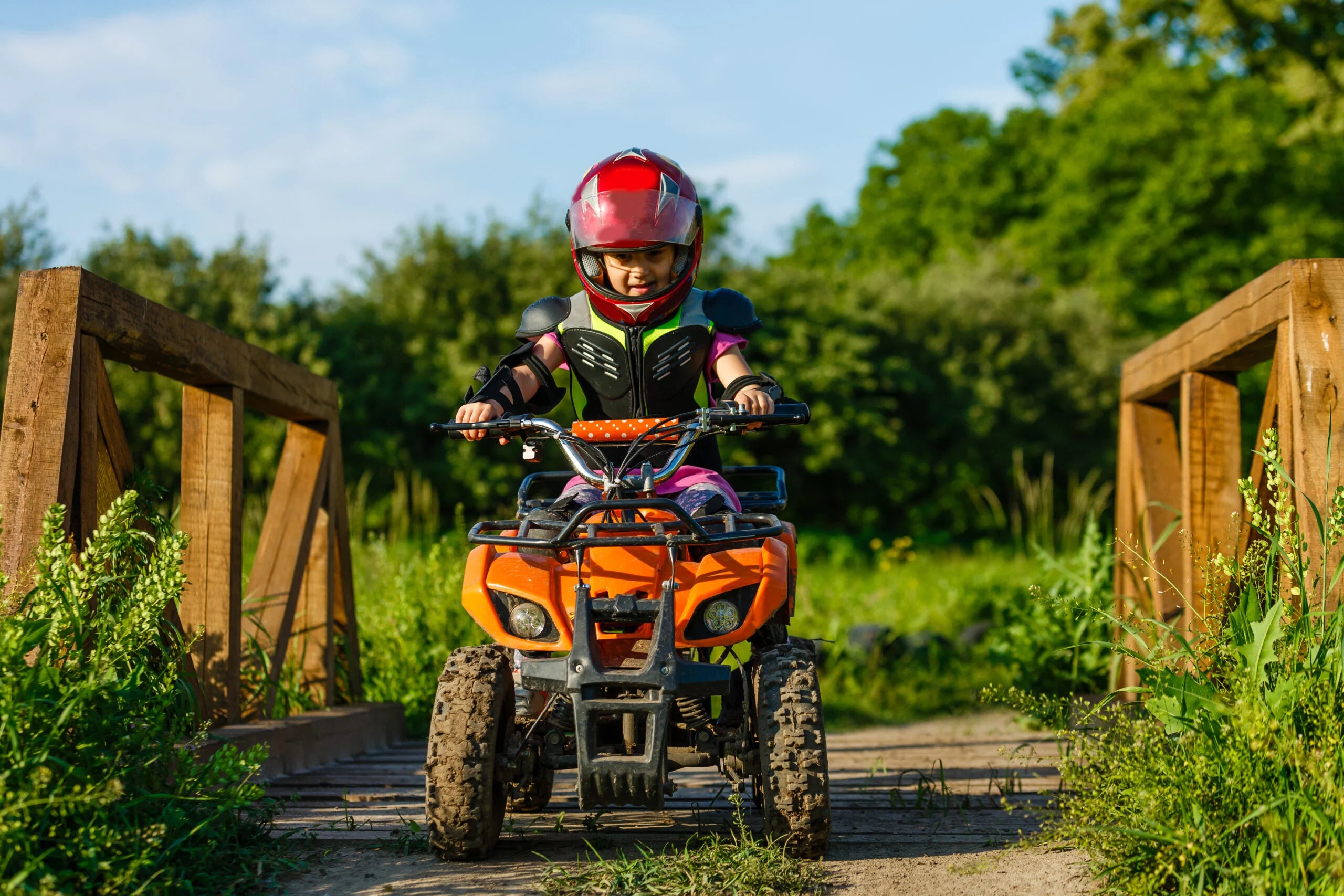 Child wearing a red helmet riding an orange ATV across a wooden bridge surrounded by greenery in daylight