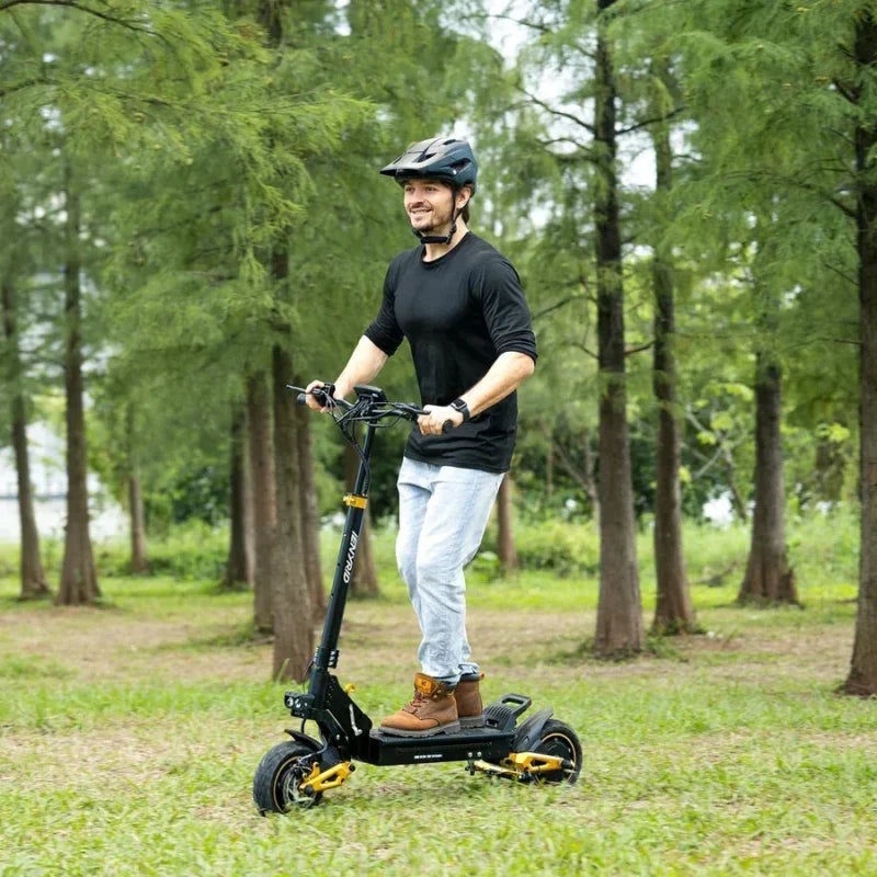 A man wearing a black helmet, black shirt, and jeans rides a black electric scooter through a grassy area with tall trees in the background.