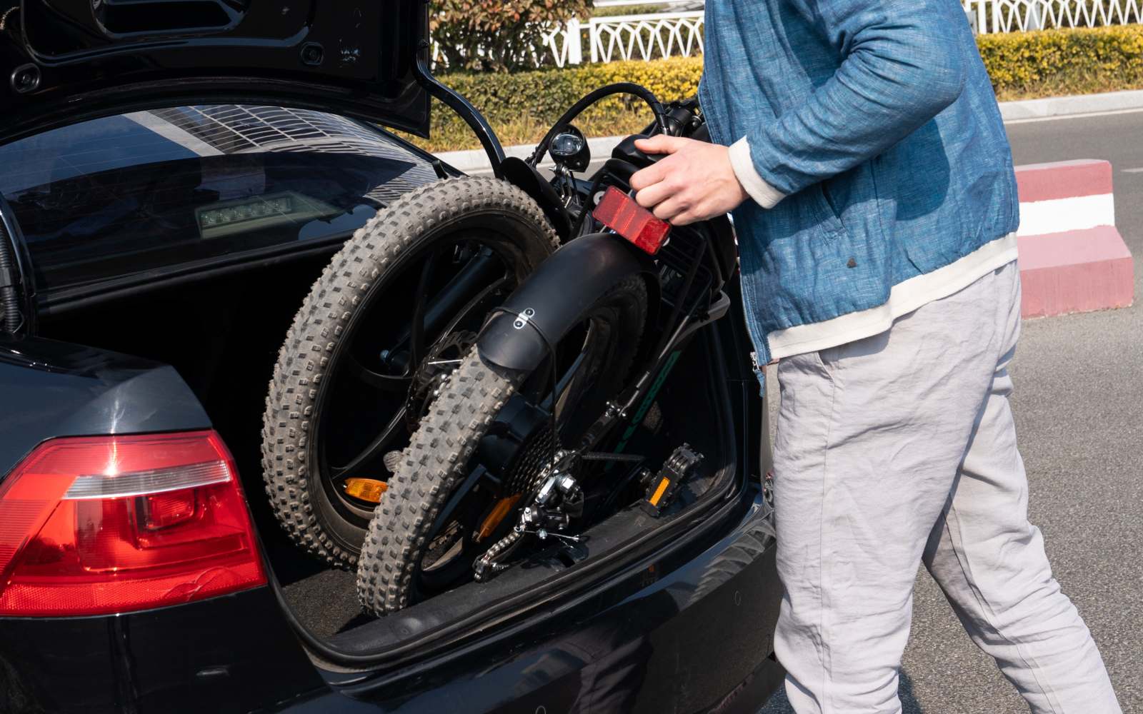A person places a folded black bicycle into the trunk of a black car on a sunny day, with part of the car and the persons upper body and arms visible.