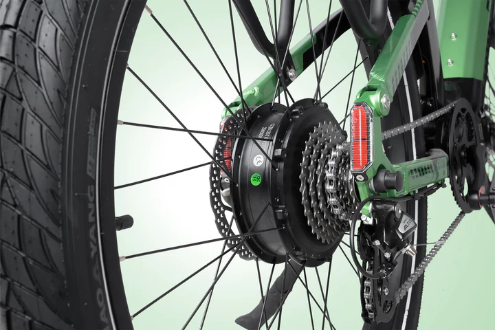 Close-up of the rear wheel of a green bicycle, showing the gears, derailleur, disc brake, and hub motor, with reflectors on the frame and spokes visible against a light background.