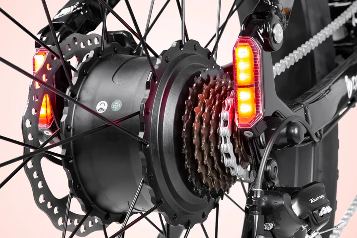 Close-up of a bicycle rear wheel showing the gear cassette, chain, disc brake, and two red LED rear lights attached to the frame near the spokes.