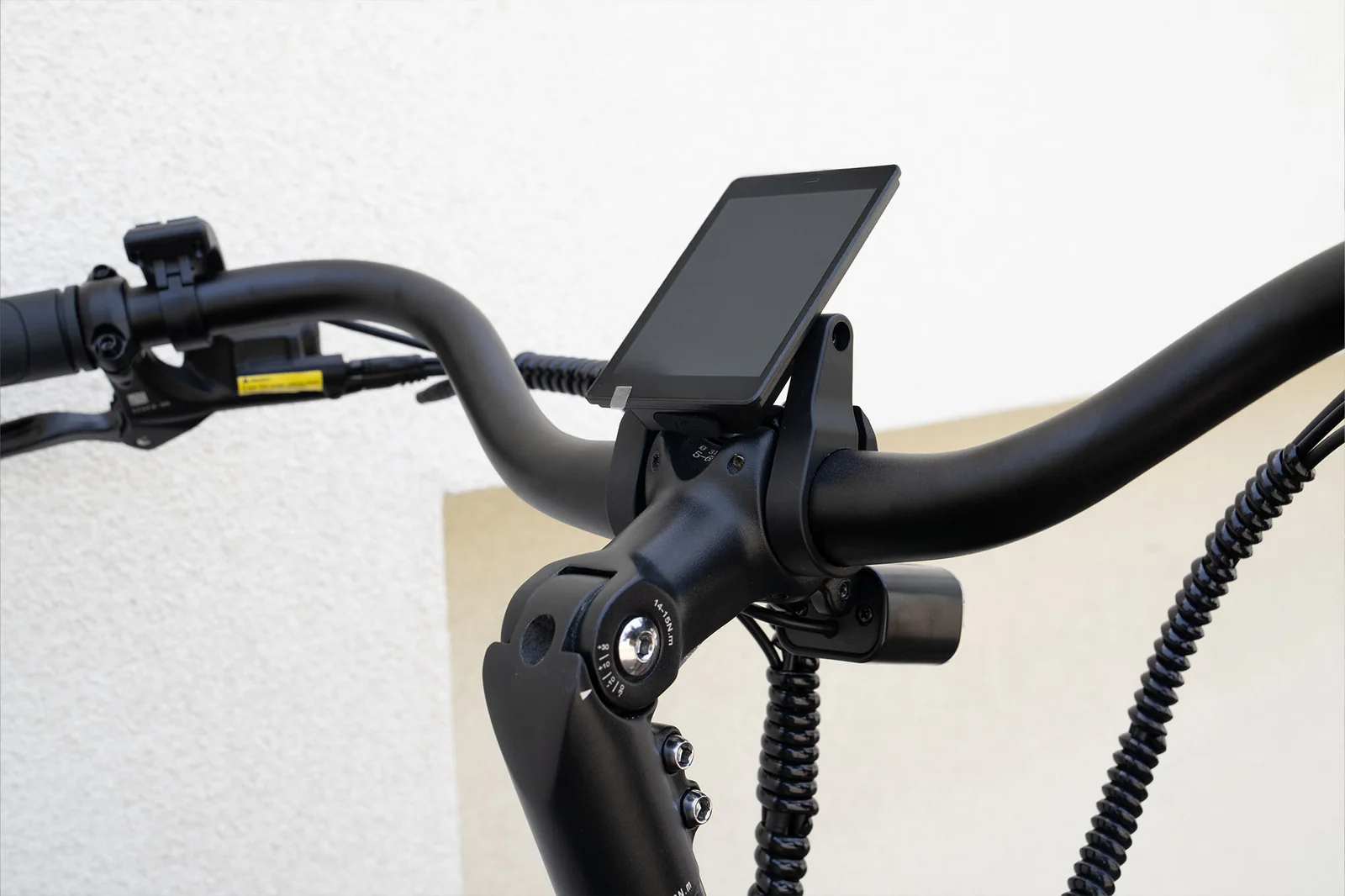 Close-up of a bicycle handlebar with a mounted digital display screen in the center. The bike has a black frame and cables, and the background is a plain, light-colored wall.