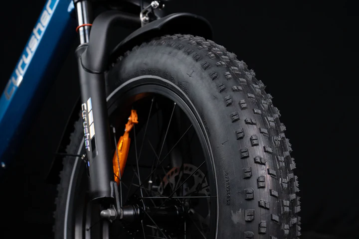 Close-up of a fat tire on an electric bicycle, showing the front wheel, thick tread, part of the blue frame, and black suspension fork against a dark background.