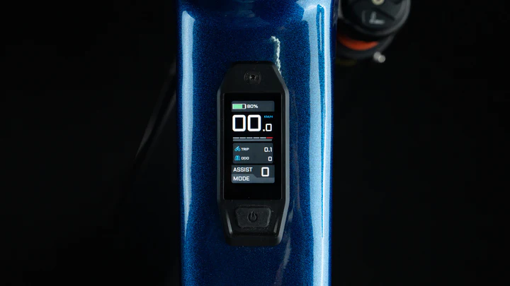 Close-up of an electronic bike display screen embedded in a blue bike frame, showing speed, battery percentage, assist mode, and other cycling data against a dark background.