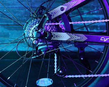 Close-up of a bicycle’s rear wheel and gear mechanism, illuminated by blue and purple lights, with a patterned frame and visible chain.
