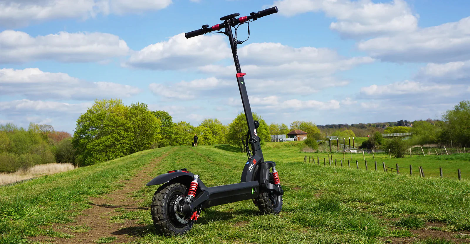 Black and red electric scooter parked on a dirt path in a green grassy field under a partly cloudy blue sky