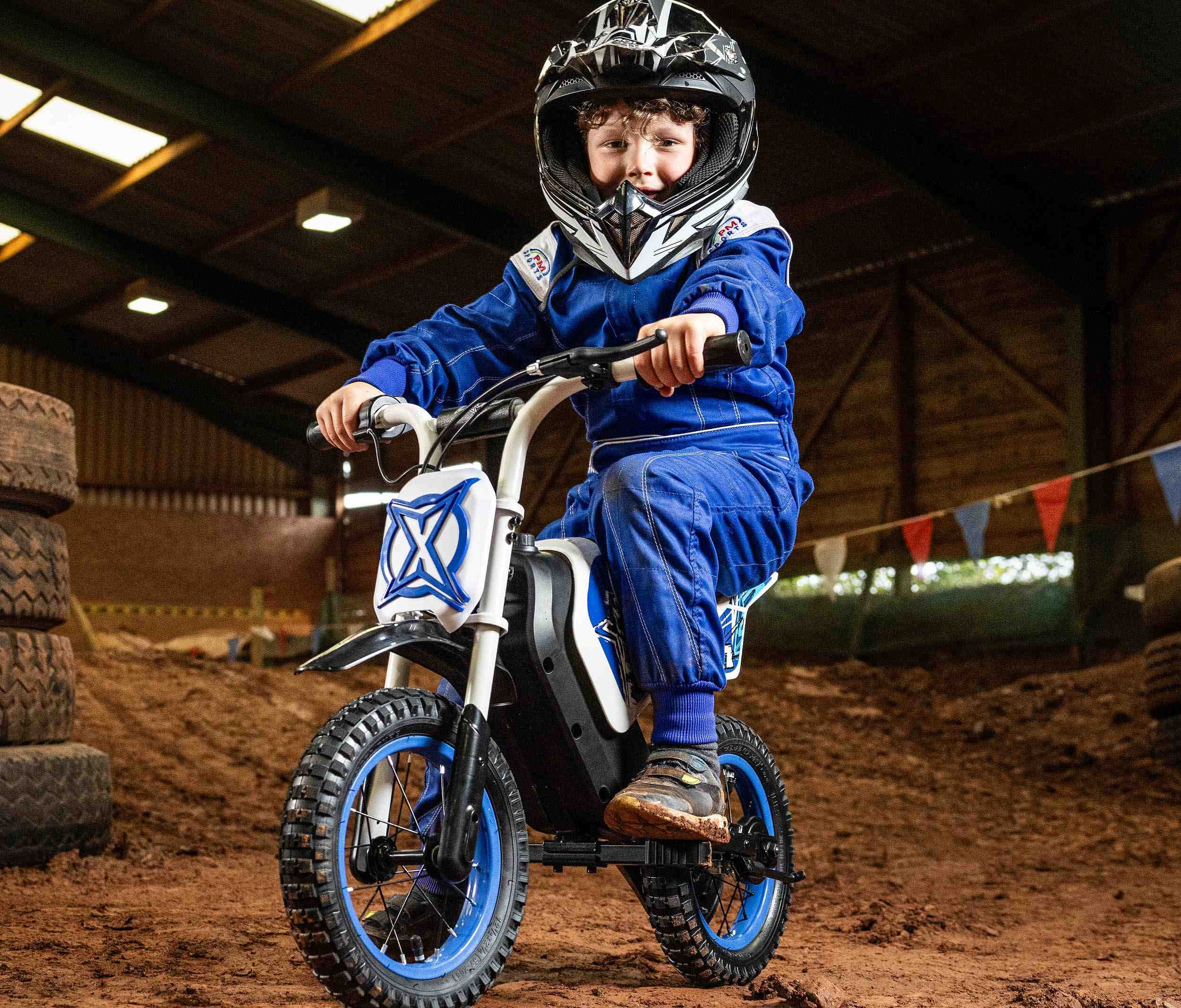 Child wearing blue racing suit and helmet riding a small electric bike indoors on dirt track