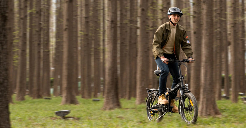 A person wearing a helmet, jacket, and boots rides a black bicycle through a forest of tall, evenly spaced trees with green grass on the ground.