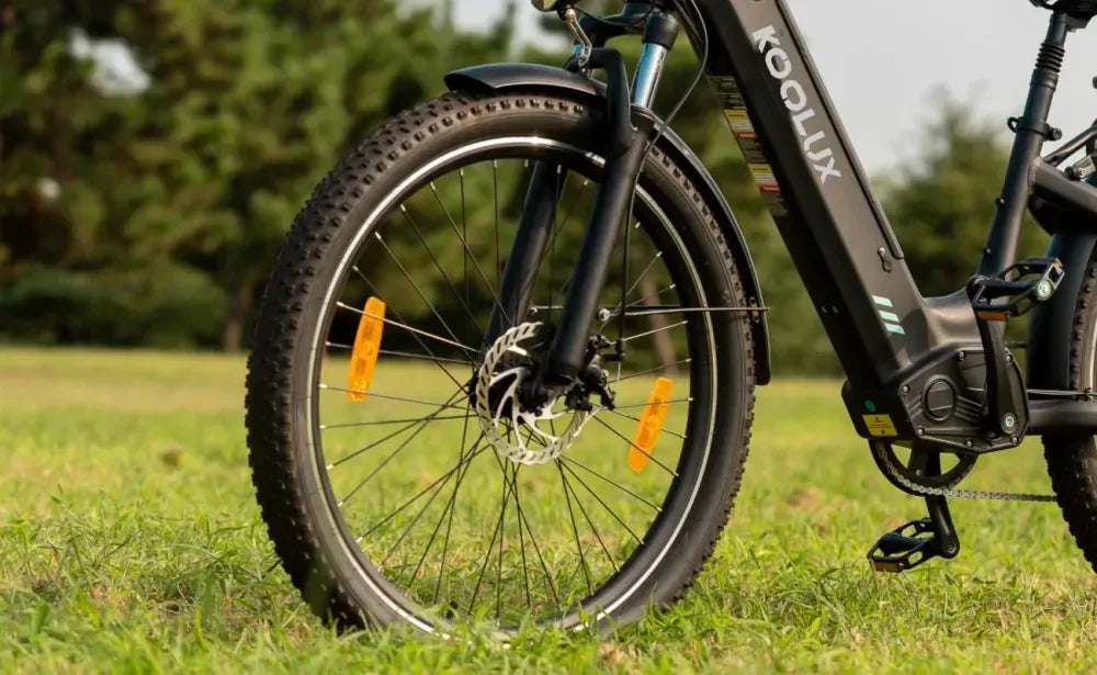 Close-up of the front wheel and pedal of a black KOO LUX electric bicycle parked on green grass, with trees blurred in the background.