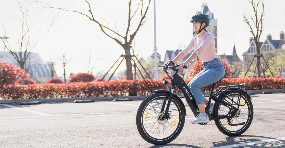 A woman wearing a helmet and casual clothes rides a black e-bike on a sunny day in an urban area, with trees, red bushes, and buildings in the background.