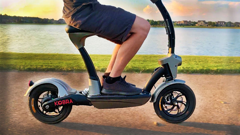 A person wearing black shorts and sneakers rides a three-wheeled electric scooter labeled KOBRA along a lakeside path, with water and houses visible in the background.
