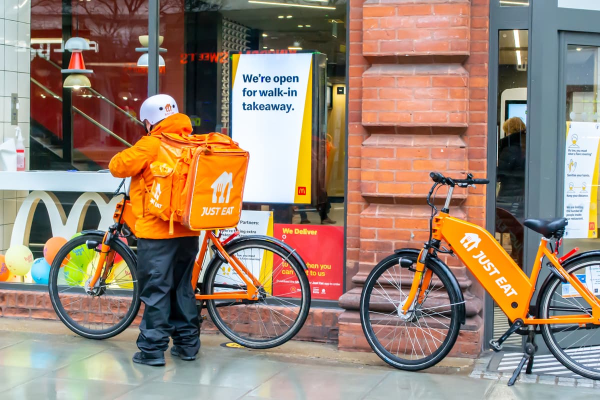 A delivery person in an orange Just Eat uniform stands beside an orange Just Eat bicycle in front of a McDonalds. A sign in the window reads, Were open for walk-in takeaway.