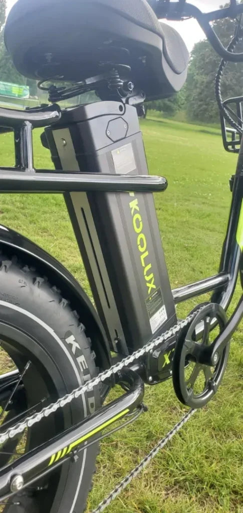 Close-up of a KOOLUX electric bike showing its battery mounted below the seat, part of the rear wheel, and the pedal, with grass and trees in the background.