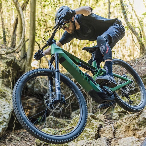 A mountain biker wearing a helmet and sunglasses rides a green bike down a rocky, forested trail, leaning forward and gripping the handlebars for control.