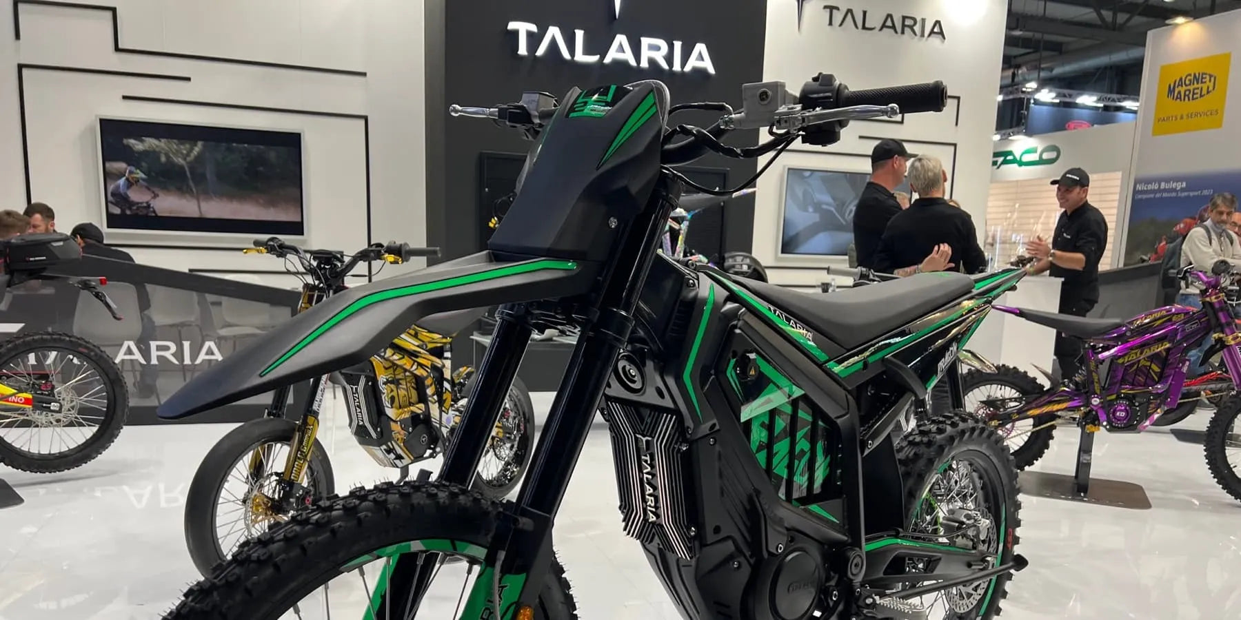 Black and green electric dirt bike displayed indoors with people and other bikes in the background at an exhibition booth.